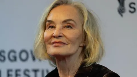 Jessica Lange in close-up at the Glasgow Film Festival. She is smiling, wearing a dark top and is in front of a white GFF banner.