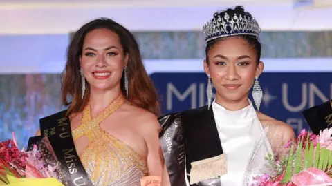 Asvin Singh Two women wearing beauty pageant sashes smile at the camera. The woman on the right is the winner Manshika Prasad, who's wearing a white dress and holding a bouquet of flowers and wearing a tiara. On the left is Nadine Roberts, wearing an orange dress and also holding a bouquet.