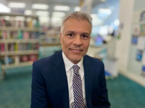 MARTIN GILES/BBC Councillor Louis Navarro looks at the camera wearing a navy suit with a white shirt. He has a spotty tie on and is smiling. He is standing in the library in Huntingdon.