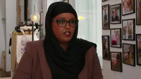 Warda Mohamed is seen in a brown blazer and black headscarf sits at a table, speaking. She wears glasses and is framed by a wall covered in photos and a coat rack in the background.