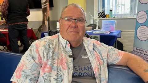 A man smiles at the camera sitting on a comfy chair. He wears a Veterans Regroup t shirt and a flowery Hawaiian style shirt over it.