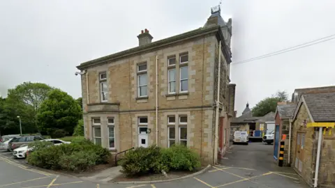 Grey skies. Light-coloured brick building with large windows with cars parked outside. There is another brick building next to it. 