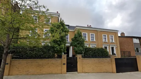 A Google Street view image of the three-storey mansion which is behind a wall with a black door and some trees