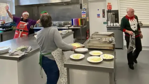 A group of volunteers working to prepare the Christmas Day lunch on Christmas Day, 2024. They can all be seen at different kitchen stations preparing food. They are all wearing Christmas themed aprons and jumpers.