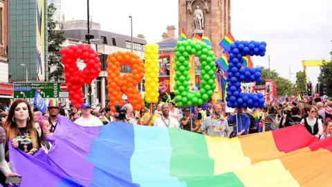 Pacemaker Large balloon letters that say Pride in rainbow colours held above a large rainbow flag.