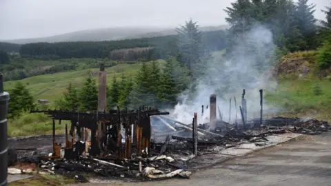 Marc Charron The smouldering remaids of a building stand on tarmac in the middle of a tree-lined country scene