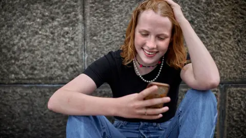 A girl looks at her phone, she has her hand on her head smiling and is wearning a black top and blue jeans. 