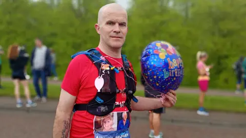 Stephen Perry, a man with a shaved head poses with a helium "happy birthday" balloon at the start of the Belfast Marathon route. He is wearing a red t-shirt and a black runner's harness containing equipment like a straw for a water pack. Other runners stand in the blurred background. 