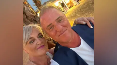 A "selfie" picture of McDonald and Mel on a beach. She has short blonde hair and is wearing pink lipstick. She has her arm around him and he is wearing a white t-shirt and blue suit waistcoat. They're both smiling.
