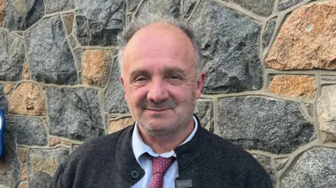 A man called Neil Inder stands in front of a stone wall. He has a dark coat on with a white shirt and red tie underneath. He is balding with short greying hair. He is unshaven.