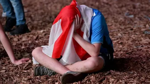 Getty Images An England fan sits on the ground with holding his head with his hands. He is hiding his face with an England flag over his head.