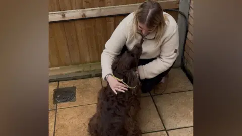 Contributed Rebecca Harvey crouches in the corner of a garden as she holds her dog flora and looks directly at her. Rebecca has long blonde hair and wears a cream jumper with dark trousers and brown shoes. Flora has brown fur and a yellow lead on. 