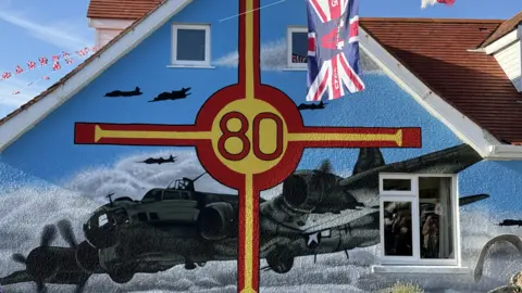 The side of a house that has the red and yellow cross of the Guernsey flag with 80 in the middle and a large painting of an American B-17 bomber.