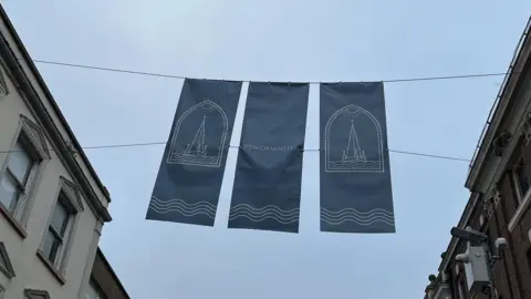 Jon Wright/BBC Three navy blue banners hanging portrait above a pedestrian street. They feature illustrated images of the tower and the words "Ipswich Minster".