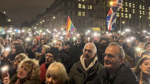 A large crowd gathered at a memorial in Parliament Square. A Pride and Union Jack flag are being waved.