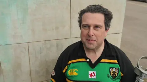 A man with swept back black hair in a green and yellow Northampton Saints shirt, sits in a wheelchair holding a crutch looking up at the camera.