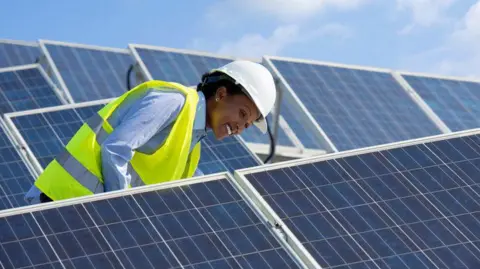 A woman engineer is checking solar photovoltaic panels at a solar farm. She is wearing a high-vis jacket and helmet and smiling.