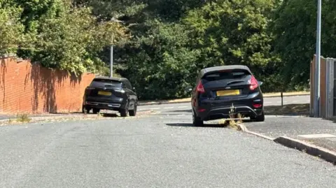 Charlie Stubbs / BBC There are two black cars, both of which are parked with half of their car on the pavement. Next to the car on the left, there is a red brick wall with green trees poking over the top of it. They are on a downwards slope.