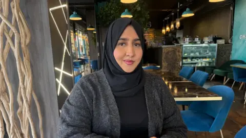 A woman wearing a black hijab and grey cardigan sits in a cafe and smiles at the camera.