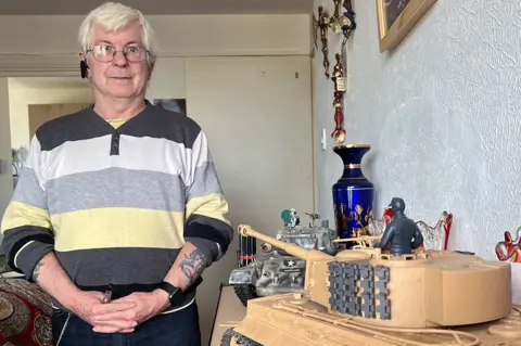 Invicta House resident David Bond stands to the left of the frame wearing a stripy coloured jumper while standing in his home next to a wall and a model tank.