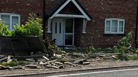 A house set back from a main road. The brick wall and fence in front of it has been destroyed, with rubble lying everywhere.