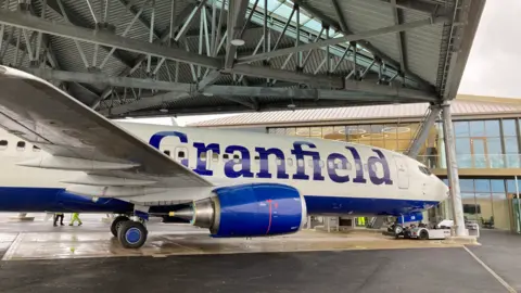 Cranfield University A white aeroplane sits under a hangar, with the word Cranfield emblazoned in blue on the side. Behind it is a modern building made mainly of glass.