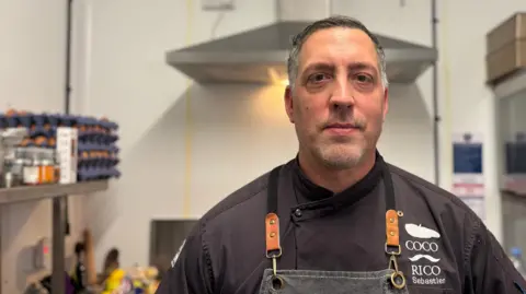 BBC Sébastien is in a commercial kitchen setting, wearing a black chef’s uniform with the name “RICO” and “COCO” embroidered on the chest. He also has a dark apron with leather straps and metal rings. Behind them are stainless steel appliances, a ventilation hood, and shelves with kitchen supplies, including a tray of eggs and flowers.
