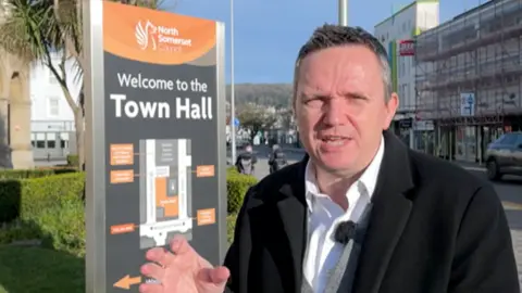 Man with short hair looking at the camera. He's stood next to a North Somerset Council Town Hall sign outside in Weston-super-Mare.