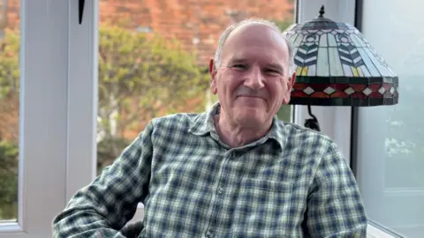 Robin Stamp Robin Stamp is seen sitting in a chair by a window with a lamp behind him. He is wearing a checked shirt and is smiling.