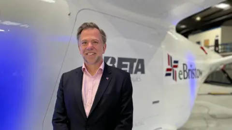 Simon Meakins stood in front of white electirc plane. He's wearing a pink shirt and black blazer. He is smiling at the camera. 