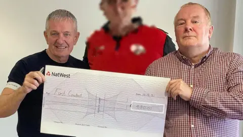 First Contact UK Mental Health Thomas Judge, left, and Thomas Carroll, right, with a blurred out man in the middle, are holding an oversized cheque for £580. Judge, at the time a man in his late 50s, is wearing a dark short sleeved top and Thomas Carroll is wearing a red checked shirt, untucked. He has short, fair hair and is in his early 60s. The man in the middle is unknown and blurred to protect his identity.