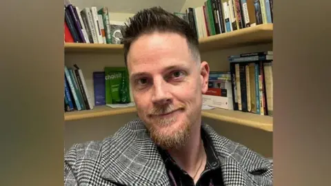 Adam Powell Dr Adam Powell, with short brown-reddish hair, with a ginger moustache and beard, wearing a black and white checked blazer, looks at the camera. A bookcase filled with books is behind him. 