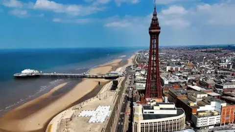 A general aerial view of Blackpool showing the tower and the town to the right, and the beach to the left with central pier. The sky is blue with a few white clouds.