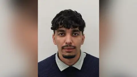 Mug shot of Zakaria Senouci, he is facing the camera against a plain light background, with dark curly hair, facial hair, and wearing a dark jumper over a light collared shirt.