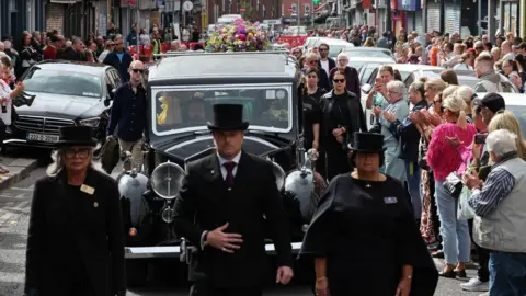 PA Media A funeral procession moves through a busy city-centre street. A black hearse is followed by a large group of people wearing black. It is led by three people wearing black clothing, two woman in dresses wearing hats and a man in a suit. 