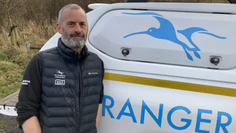 Gary Pickles is standing next to a van with the word ranger on it. He is wearing a black jacket with the Northumberland National Park logo on it and has grey hair and a grey beard. He is a man in his 40s or 50s.