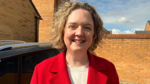 Anna Sabine stands outside on a sunny day and poses for a head and shoulders photo. She is wearing a red coat and a white T-shirt and has short, curly blonde hair. She is standing in front of a red brick wall.