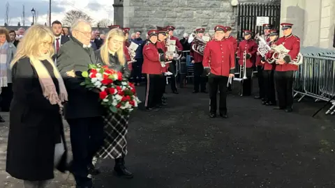 A man is holding a wreath. He's being supported by two women. A band are wearing maroon uniforms and holding instruments. A crowd of people are watching. 