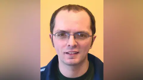 Pacemaker A man is standing in front of a yellow wall. He has dark, receding hair and glasses. He is wearing a green t-shirt and navy fleece. 