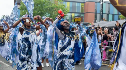 The troupe wears blue and white garments, their faces are painted and their arms raised in the air. They pass a crowd of onlookers standing behind metal barriers.