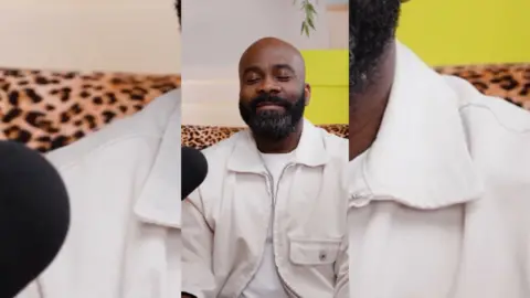 A man, Melvin Odoom, with a bread, wearing a beige jacket over a white t-shirt. He is smiling with his eyes closed.