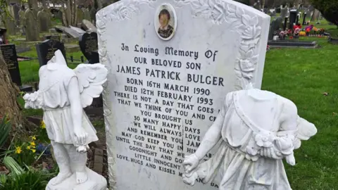 Statues of two cherubs with missing heads stand next to a gravestone for James Bulger.