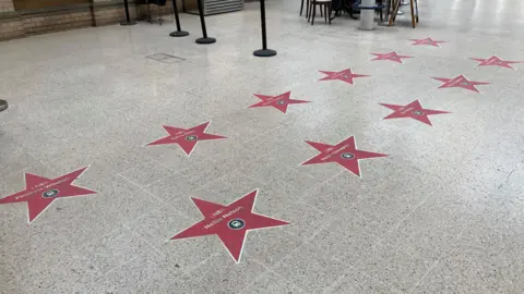 A large indoor public space with a smooth, light-coloured floor. Scattered across the floor are multiple large red stars, similar in style to “Hollywood Walk of Fame” stars. Each star has a white border and a circular emblem in the centre.