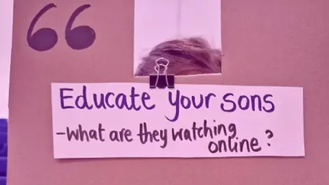 A cardboard sign is held up to the camera, in marker pen it says 'Educate your sons - what are they watching online?' 