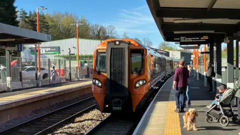 A train pulling into Kings Heath Railway Station. It is black and orange and has at least three carriages. There are people standing on the platform, as well as a brown dog and a pushchair
