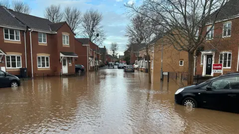 BBC A flooded residential street in Taunton. Brown water can be seen flooding the road, reaching half-way up the tyres of cars parked by the red-brick houses.