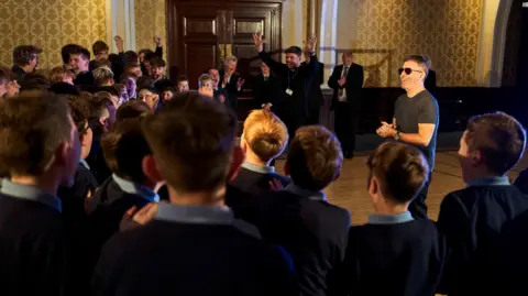 BGT/TalkbackThames/ITV Simon Cowell stands to the right of the boys who are in a semi circle gathered around him laughing and cheering, some seen from the back. They were navy school jumpers and light blue polo shirts