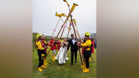 Ian Foster Amber is wearing a white wedding dress and Alex Richardson is dressed in a black jacket and trousers, white shirt and black bow tie.
They are smiling and Amber is looking up at yellow wellies held aloft on boat oars as they walk through Looe RNLI’s yellow welly guard of honour. Crew members are wearing drysuits and red lifejackets and yellow helmets. 