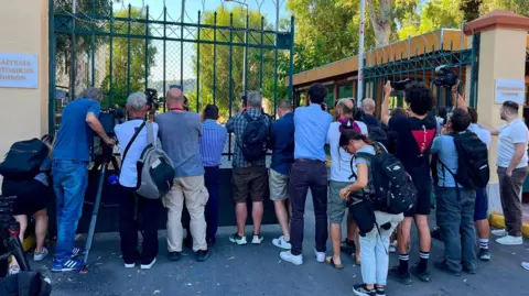 A group of journalists and camera crews gather outside a tall metal security gate, aiming cameras and recording equipment through the fence toward a location inside the compound. Signs with Greek text are mounted on the walls beside the entrance. Trees and buildings are visible beyond the gate, and the scene takes place in bright daylight on a city street.