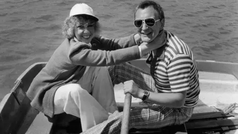 Hulton Archive A black and white image of Parkin with her husband Patrick Hughes, they are sat on a rowing boat. Hughes has his left arm holding an oar. He is wearing a horizontal striped t-shirt and checked trousers. He is wearing sunglasses. Parkin is wearing white trousers and a cardigan, her curly, short hair is covered with a white hat. She has her arms around Hughes' throat in a jokey fashion.